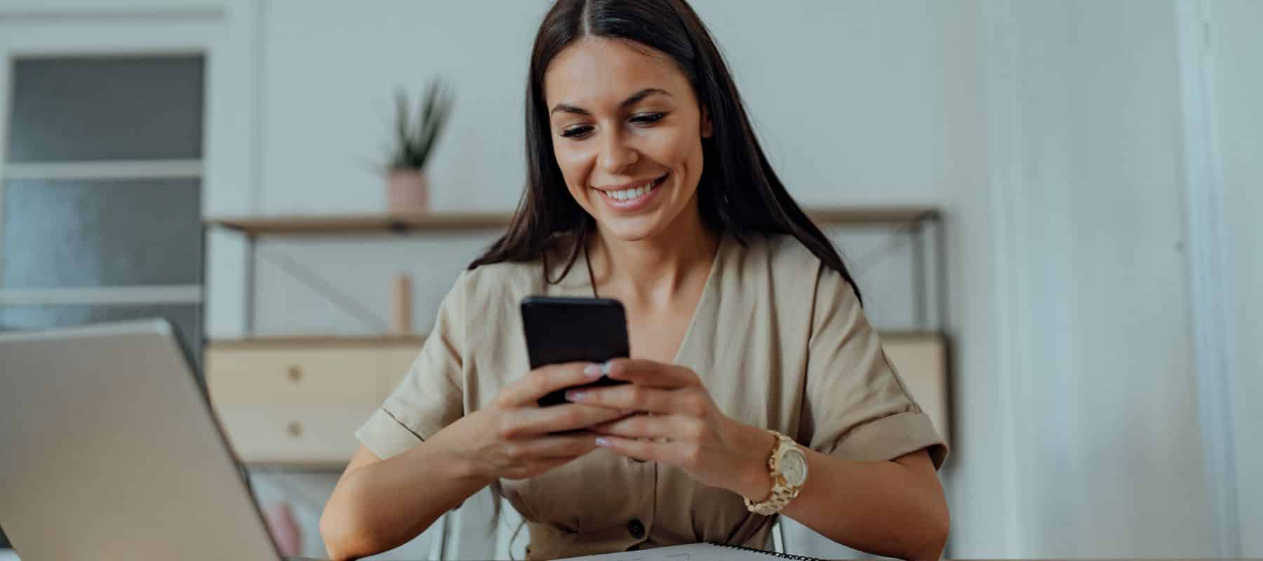 A woman sits at a desk smiling while using her smartphone, with a laptop and shelves in the background
